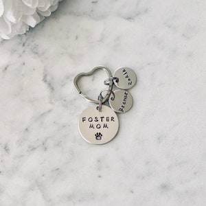 May include: Silver keyring with a heart-shaped ring and three round charms. The largest charm says "Foster Mom" with a paw print. The other two charms say "Brewer" and "Rosie".