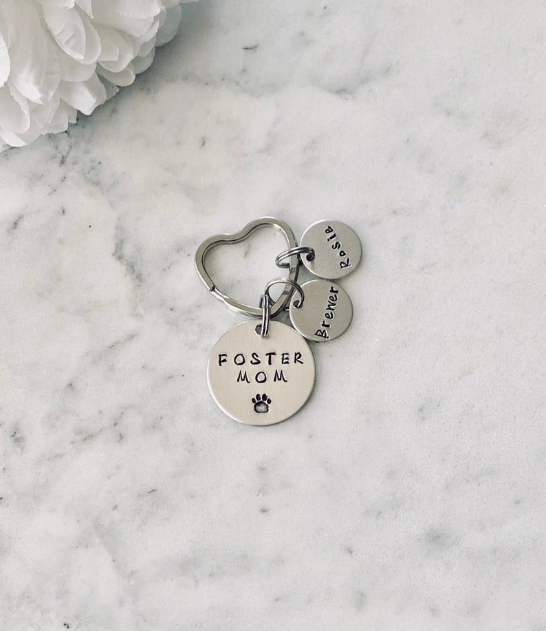 May include: Silver keyring with a heart-shaped ring and three round charms. The largest charm says "Foster Mom" with a paw print. The other two charms say "Brewer" and "Rosie".