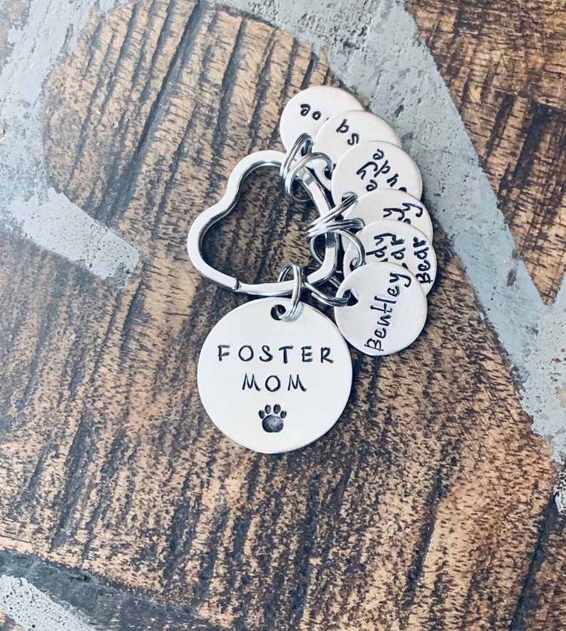 May include: Silver keyring with a heart-shaped ring and multiple round tags. The largest tag says "Foster Mom" with a paw print. The other tags have names written on them: "Bear", "Bentley", "Andy", "Ava", "Zoe", and "Bo".