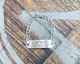 Puede incluir: Un brazalete de cadena de plata con una etiqueta rectangular grabada con las coordenadas "N 42° 59' 9" W 85° 39' 42" ".