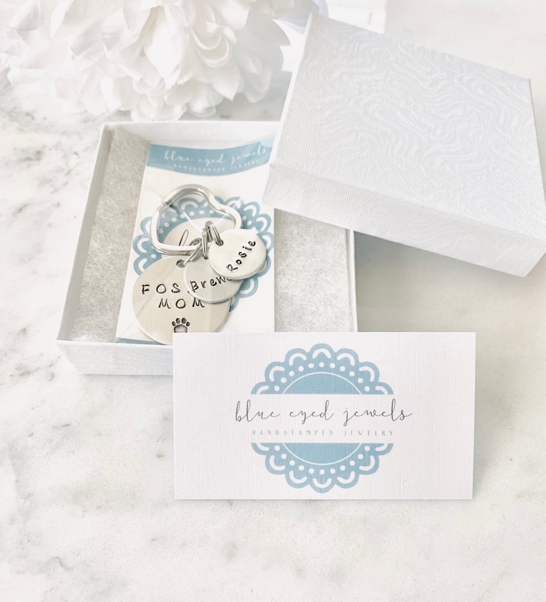 May include: A silver keychain with three charms. The charms are round and have the words "Rosie", "Brew", and "Fos Mom" engraved on them. The keychain is in a white box with a blue and white card that says "blue eyed jewels handstamped jewellery".