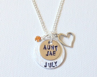 Puede incluir: Collar plateado con múltiples colgantes. El collar presenta una pequeña gema naranja, un colgante en forma de corazón y dos colgantes circulares. El colgante superior dice "AUNT JAE" y el colgante inferior dice "JULY".