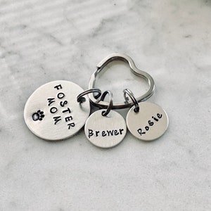 May include: Silver heart-shaped keyring with three round tags. The largest tag says "Foster Mom" with a paw print. The other two tags say "Brewer" and "Rosie".