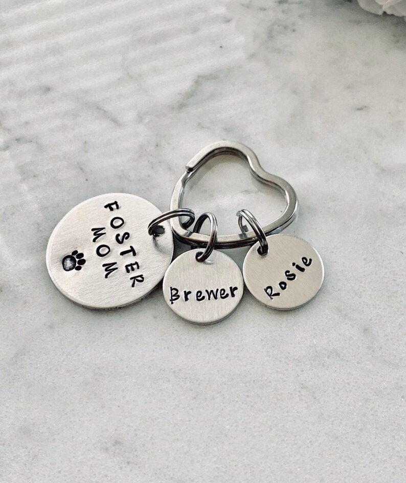 May include: Silver heart-shaped keyring with three round tags. The largest tag says "Foster Mom" with a paw print. The other two tags say "Brewer" and "Rosie".