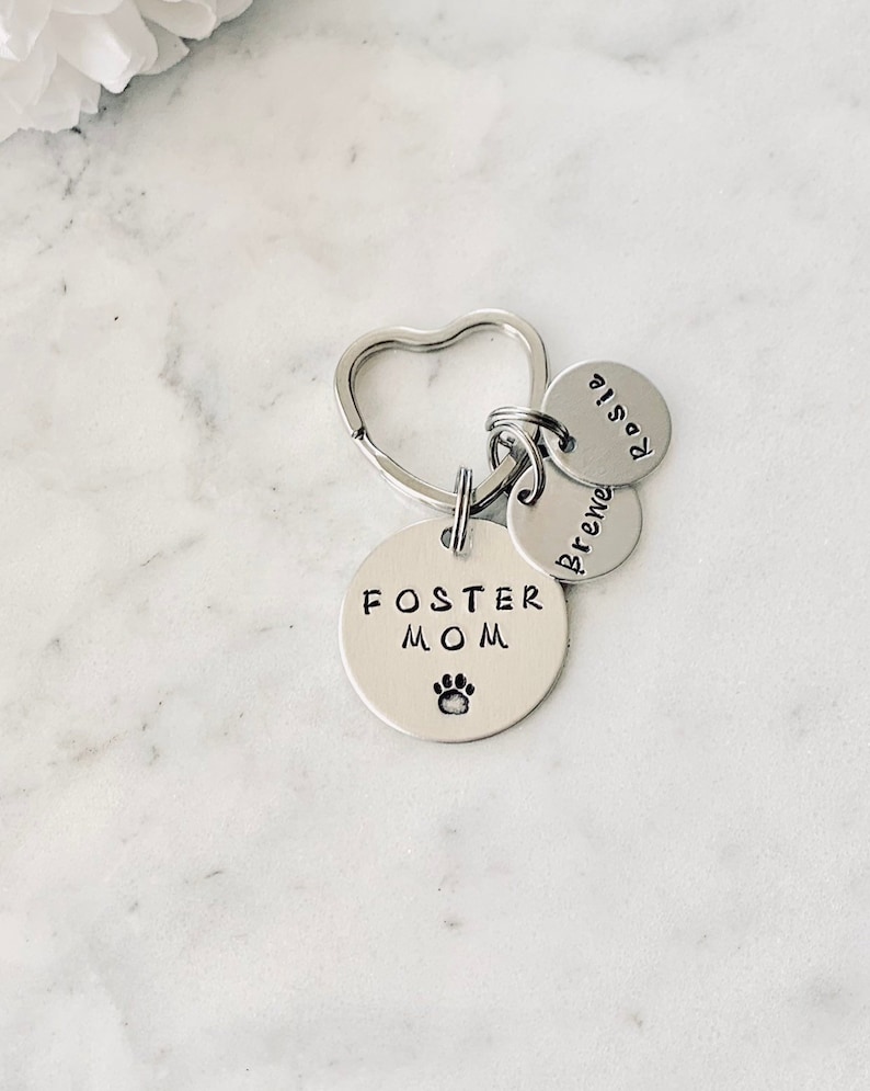 May include: Silver keyring with a heart-shaped ring and three round charms. The largest charm says "Foster Mom" with a paw print. The other two charms say "Brenda" and "Rosie".