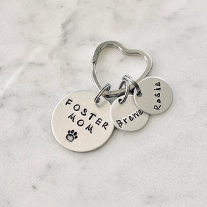 May include: Silver keychain with three tags. The largest tag says "Foster Mom" with a paw print. The other two tags say "Rosie" and "Brewe".