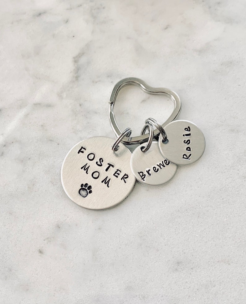 May include: Silver keychain with three tags. The largest tag says "Foster Mom" with a paw print. The other two tags say "Rosie" and "Brewe".