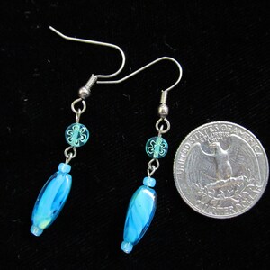 May include: A pair of dangle earrings with silver-tone hooks. Each earring features a small, round, light blue bead with a floral design, a small silver bead, and a larger, oval-shaped, blue glass bead with white swirls. A quarter is next to the earrings for size comparison.