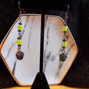May include: A pair of dangle earrings with silver-tone hooks. Each earring features a series of lime green beads, small dark beads, and a dark purple bead with a decorative metal cap. The earrings are displayed on a black stand.