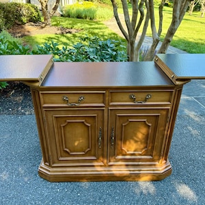 May include: A wooden buffet cabinet featuring a dark brown top with two extendable side leaves. The front includes two drawers with brass-toned metal pulls and two lower cabinet doors with matching hardware and recessed rectangular panel detailing.