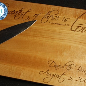 May include: A wooden cutting board with engraved text. The text reads "The greatest of these is love" and "David & Briana August 5, 2006". A knife and a tomato are on the board.