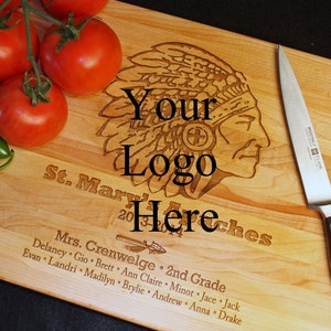 May include: A wooden cutting board with a Native American headdress design, the text "Your Logo Here", and the text "St. Mary's 2nd Grade" with a list of names. The board is light brown and has a natural wood grain. A silver knife with a black handle is lying on the board.