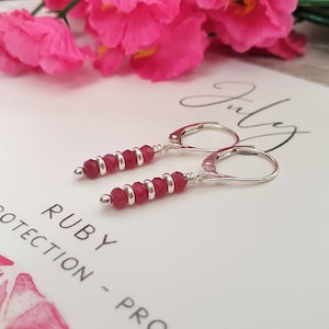 May include: Silver earrings with red ruby gemstone beads. The earrings are on a white card with the text "RUBY - PASSION - PROTECTION - PROSPERITY" and the word "July" written in cursive.