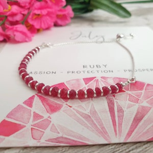 May include: A silver chain bracelet with red ruby beads. The bracelet has an adjustable sliding clasp. The card behind the bracelet says "Ruby - Passion - Protection - Prosper".