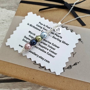 May include: A silver necklace with a pea pod charm made of wire and freshwater pearls. The pearls are in the colours of birthstones for each month of the year. The card reads "Pea in Pod Using Swarovski Pearls - Sterling Silver" and lists the birthstone colours for each month.