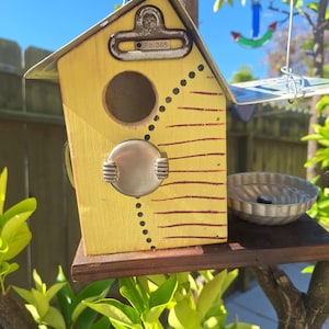 May include: A yellow wooden birdhouse with a metal roof and a circular entrance hole. It features a metal clipboard clip at the top labeled No. 365, a round silver metal accent, horizontal brown lines, and a small metal bowl attached to the base.