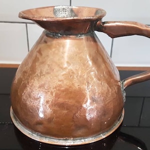 May include: A copper jug with a wide base and a curved handle. The jug has a spout and a hammered texture, giving it a rustic appearance. The copper has a warm, reddish-brown colour. The jug is likely used for serving beverages.