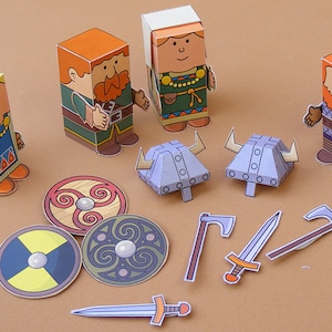Könnte beinhalten: Ein Set aus Papierfiguren von Wikingern, darunter eine Frau, zwei Männer mit Bärten und zwei Helme. Die Figuren sind bunt und detailliert gestaltet. Das Set enthält außerdem zwei Schilde, zwei Schwerter und eine Axt.