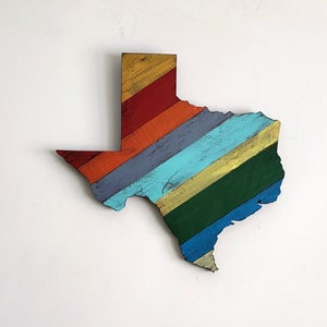 May include: A wooden cutout of the state of Texas painted in a rainbow pattern. The colors are red, orange, yellow, green, blue, and purple.