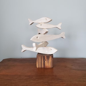 May include: A sculpture of six wooden fish, painted white with a distressed finish, stacked on top of each other. The base of the sculpture is a small, square wooden block. The fish are of varying sizes.