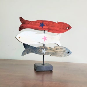 May include: A wooden sculpture of three fish stacked on top of each other. The fish are painted in red, white, and blue with stars on each. The sculpture is on a black wooden base.