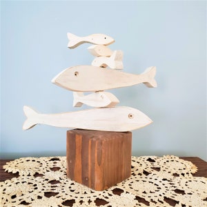 May include: A stack of five white wooden fish sculptures, each with a slightly different shape and size, are balanced on a brown wooden block. The fish are arranged in a pyramid shape, with the largest fish at the bottom and the smallest fish at the top.