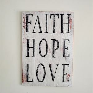 May include: White wooden sign with distressed paint and black lettering that reads "FAITH HOPE LOVE".
