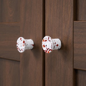 Può includere: Due pomelli circolari per armadietto con base bianca e un motivo maculato composto da frammenti rossi, arancioni e neri. I pomelli sono montati su un'anta di armadietto in legno marrone scuro con venature verticali e un pannello orizzontale.