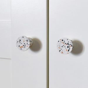 以下が含まれることがあります： 白いキャビネットドア2枚のクローズアップ。丸い、斑点のあるキャビネットノブが付いています。ノブは、黒、オレンジ、白の斑点模様のテラゾーのようなデザインです。ドアは、くぼんだパネルデザインのプレーンホワイトです。