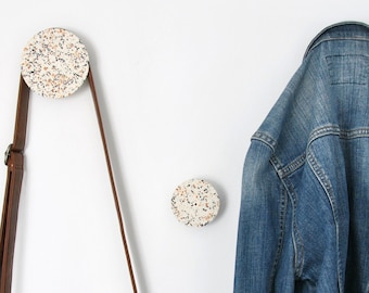 May include: Two circular wall hooks featuring a speckled terrazzo pattern in shades of white, gray, and orange. A brown leather crossbody bag hangs from the left hook, and a blue denim jacket hangs from the right hook on the plain white wall.