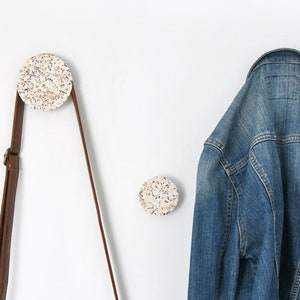 Op de afbeelding: Twee ronde wandhaken met een gespikkeld terrazzopatroon in tinten wit, grijs en oranje. Een bruine leren schoudertas hangt aan de linkerhaak en een blauw spijkerjack hangt aan de rechterhaak aan de effen witte muur.
