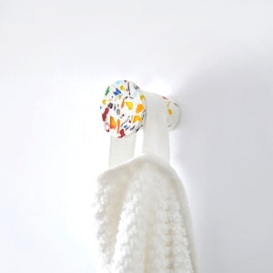 Può includere: Un gancio bianco a parete con una testa circolare decorata con schegge colorate in stile terrazzo. Un asciugamano bianco è appeso al gancio. Il gancio è progettato per appendere asciugamani, accappatoi o altri oggetti.