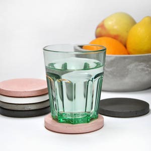 Puede incluir: Un vaso de agua sobre un posavasos de hormig&oacute;n rosa. El posavasos est&aacute; encima de una pila de otros cuatro posavasos en negro, gris, blanco y rosa. Hay fruta en un cuenco en el fondo.
