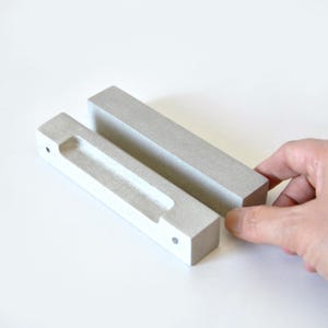 Peut inclure: Deux poignées de tiroir rectangulaires en béton gris clair. L'une a une poignée encastrée, l'autre est pleine. Les poignées de tiroir sont présentées sur une surface blanche. Le béton a une texture mouchetée.