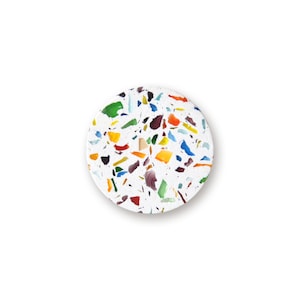 May include: A circular coaster featuring a white base embedded with a scattered pattern of multi-colored glass fragments. The surface displays various irregular shapes in shades of green, blue, yellow, orange, red, and dark purple throughout.