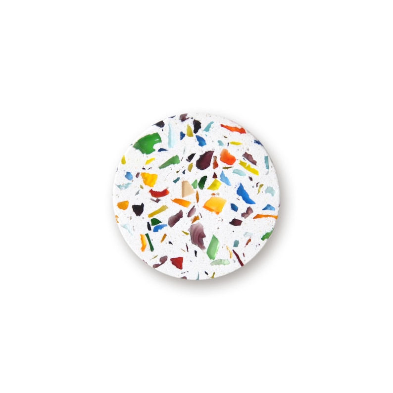May include: A circular coaster featuring a white base embedded with a scattered pattern of multi-colored glass fragments. The surface displays various irregular shapes in shades of green, blue, yellow, orange, red, and dark purple throughout.
