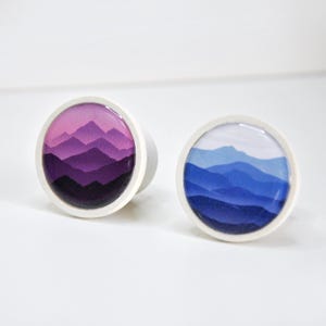 Peut inclure: Deux boutons de meuble ronds avec des motifs de montagnes. L'un présente un dégradé de tons violets, l'autre un dégradé de bleus. Les deux boutons ont un bord blanc et une base cylindrique.