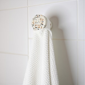 Peut inclure: Une serviette blanche est accrochée à un crochet rond en terrazzo. Le crochet est fixé à un mur carrelé blanc.