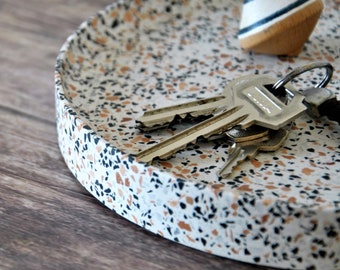 May include: A round, white tray with a speckled terrazzo pattern in black, brown, and beige. The tray has a raised rim and is filled with keys and a wooden spinning top.