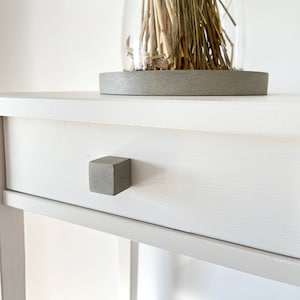 Op de afbeelding: Witte nachtkastje met een lade en een betonnen ladegreep. De lade is gesloten. Het nachtkastje staat op een wit oppervlak.