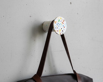 May include: A cylindrical wall hook featuring a white base with a multicolored terrazzo pattern on the circular front face. The hook is mounted to a wall and supports a dark gray bag with brown leather straps hanging from its rounded surface.