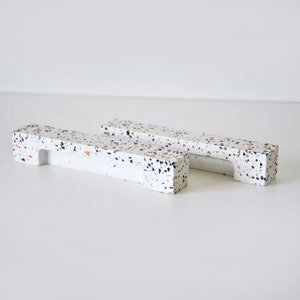 Könnte beinhalten: Zwei weiße Terrazzo-Schubladengriffe in rechteckiger Form mit einem ausgeschnittenen Griff. Die Griffe haben ein gesprenkeltes Muster mit schwarzen, braunen und weißen Flecken.