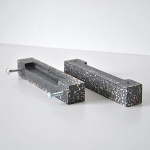 Peut inclure: Deux poignées de tiroir en terrazzo noir et blanc de forme rectangulaire avec une poignée encastrée. Les poignées ont un motif moucheté et sont fixées avec des vis argentées.