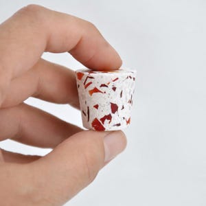 May include: A small, truncated cone-shaped knob featuring a white base embedded with scattered, irregular fragments of deep red and orange material, creating a speckled terrazzo-like appearance across the entire exterior surface of the object.