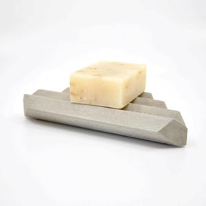 Könnte beinhalten: Ein hellgrauer Seifenhalter aus Beton mit geometrischem Design, der ein rechteckiges Stück beige Seife hält. Der Seifenhalter hat ein abgestuftes Design für die Entwässerung. Die Seife hat sichtbare Flecken.