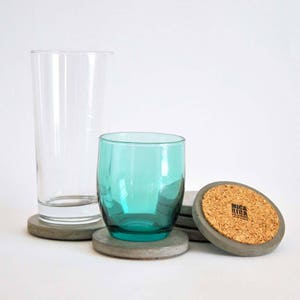 Puede incluir: Tres posavasos de hormig&oacute;n gris con una parte superior de corcho. La parte superior de corcho tiene el texto "BRICK RIG" impreso. Dos vasos de vidrio transparente y un vaso de vidrio turquesa est&aacute;n sobre los posavasos.