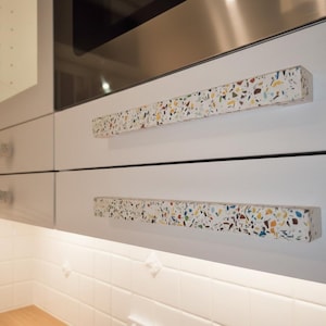 May include: Two long, rectangular cabinet handles featuring a white base with a multi-colored terrazzo pattern of blue, yellow, and brown flecks. The handles are mounted horizontally on smooth, matte white kitchen drawers below a stainless steel appliance.