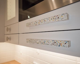 Puede incluir: Dos tiradores de armario largos y rectangulares con una base blanca y un patrón de terrazo multicolor con motas azules, amarillas y marrones. Los tiradores están montados horizontalmente en cajones de cocina blancos mate y lisos.