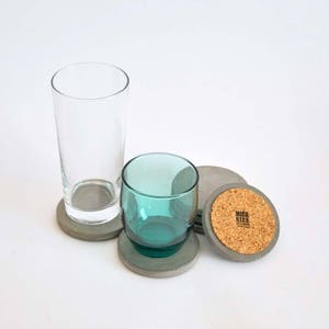 Puede incluir: Conjunto de cuatro posavasos de hormig&oacute;n gris con parte superior de corcho. Los posavasos son redondos y tienen una superficie lisa. Un posavasos tiene el texto "MICA RIGA" impreso en el corcho.