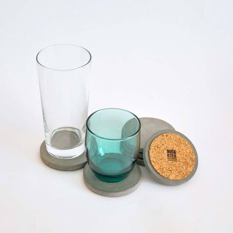 Puede incluir: Conjunto de cuatro posavasos de hormig&oacute;n gris con parte superior de corcho. Los posavasos son redondos y tienen una superficie lisa. Un posavasos tiene el texto "MICA RIGA" impreso en el corcho.